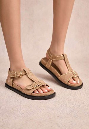 Pieds portant des sandales en daim beige avec des boucles et des brides cloutées sur un sol clair texturé.