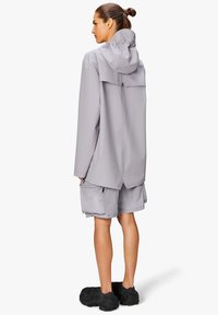 Jasnoszara wodoodporna kurtka z kapturem, luźny krój, plisowany tył, połączona z szarymi szortami i teksturowanym czarnym obuwiem.