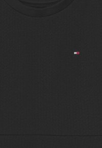 Czarny sweter o teksturowanej fakturze z prążkowanym okrągłym dekoltem. Na piersi ma małe haftowane logo w kolorach czerwonym, białym i niebieskim.