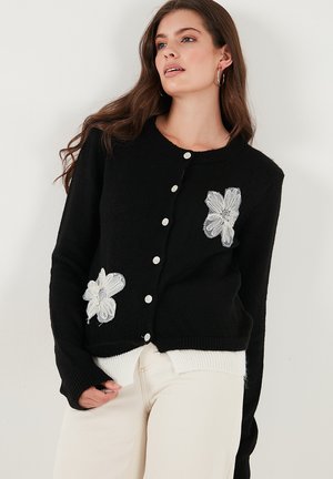 Black cardigan with white flower embroidery, round neckline, and white buttons. Knit texture with ribbed cuffs and hem. Layered white bottom.