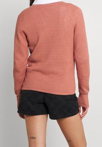Rosafarbener Strickpullover mit strukturiertem Muster, enganliegenden Bündchen und weitem Ausschnitt, kombiniert mit schwarzen karrierten Shorts.