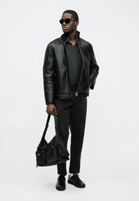 Schwarze Lederjacke mit Reißverschluss, dunkelgraues Strickoberteil, schwarze Hose, elegante schwarze Sneakers und eine strukturierte schwarze Umhängetasche.