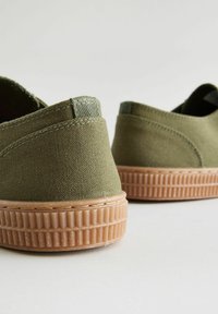 Baskets slip-on en toile verte avec des semelles en caoutchouc texturé couleur beige et des détails de couture subtils, présentant un design discret et une finition soignée.