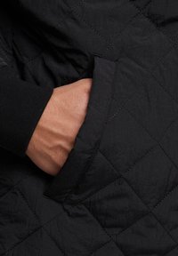 Giacca nera trapuntata con una mano in una tasca laterale. Il tessuto appare liscio con una leggera lucentezza e presenta un motivo a diamante. Dettaglio del polsino nero visibile.