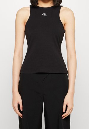 Kvinde iført en sort, ærmeløs Calvin Klein tanktop og sorte bukser, stående med armene afslappet ned langs siden.