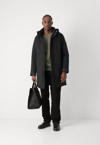Schwarze gepolsterte Jacke mit Kapuze, grüner Pullover, schwarze Hose und schwarze Schuhe; hält eine strukturierte schwarze Tasche mit texturiertem Design.