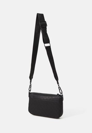 Sort crossbody taske med justerbar bred rem og præget mønster, hængende mod en ensartet lys baggrund.