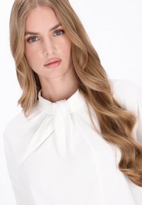 Blusa bianca con dettaglio al collo annodato, tessuto morbido, caduta fluida e capelli lunghi e ondulati che incorniciano il viso su uno sfondo neutro.