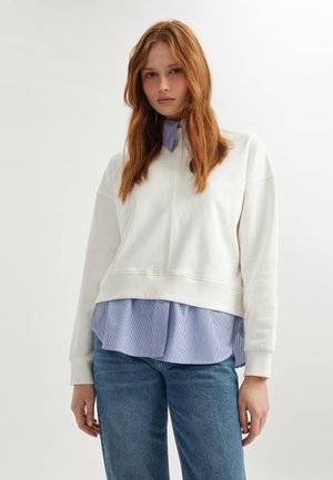 Sweatshirt blanc avec un col rond et un design à épaule tombante, superposé sur une chemise rayée bleu clair, associé à un jean en denim bleu.