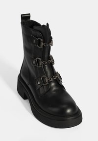 Bottine en cuir noir avec chaînes en métal décoratives, accents argentés et semelle épaisse. Comprend une languette de tirage et un design structuré.