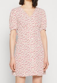 Kvinna i en vit kortärmad klänning med röd blommönster och V-ringning, som visar överkroppen och är i midjehöjd.