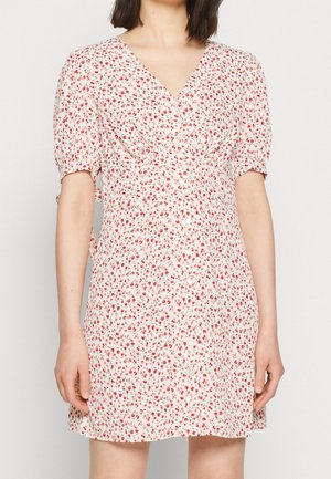 Kvinde iført en hvid kortærmet kjole med rødt blomsterprint og V-hals, der viser overkroppen og går til midten af låret.