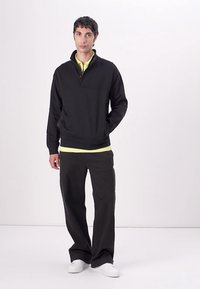 Pullover nero con zip sopra una maglietta verde lime, abbinato a pantaloni neri e scarpe da ginnastica bianche. Tessuto liscio, vestibilità rilassata e design minimale.