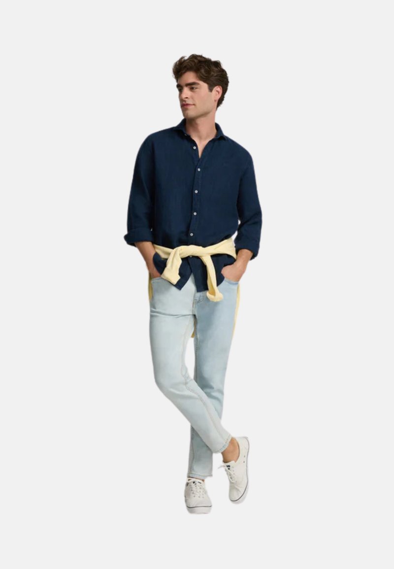 Camisa de botones azul marino, atada a la cintura con un cinturón amarillo claro, combinada con jeans azul claro y zapatillas blancas.