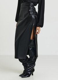 Jupe en cuir noir avec une fente et des détails froncés sur le côté, associée à des bottes en cuir noir arrivant jusqu'au genou, à bouts pointus et texture décontractée.