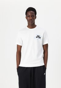Bílý bavlněný tričko s černým a modrým logem Nike Air na hrudi, kulatým výstřihem a krátkými rukávy. V kombinaci s tmavými kalhotami.