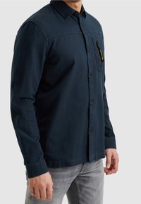 Homme portant une chemise bleu foncé à boutons avec manches longues et une poche poitrine zippée, associée à un jean gris clair.