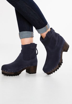 Bottes chelsea en daim marine avec semelles crantées et talons compensés en bois, portées avec un jean denim foncé retroussé.