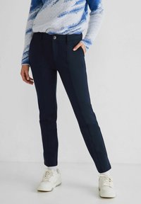 Pantaloni sartoriali blu navy a vestibilità slim, con dettagli di cucitura visibili e chiusura anteriore con bottone, abbinati a sneaker bianche.