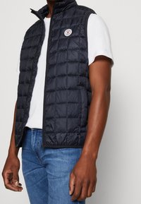 Marinblå vadderad väst med hög krage och dragkedja, med en rund logotyp-patch. Bärs över en vit t-shirt och matchas med blå jeans.