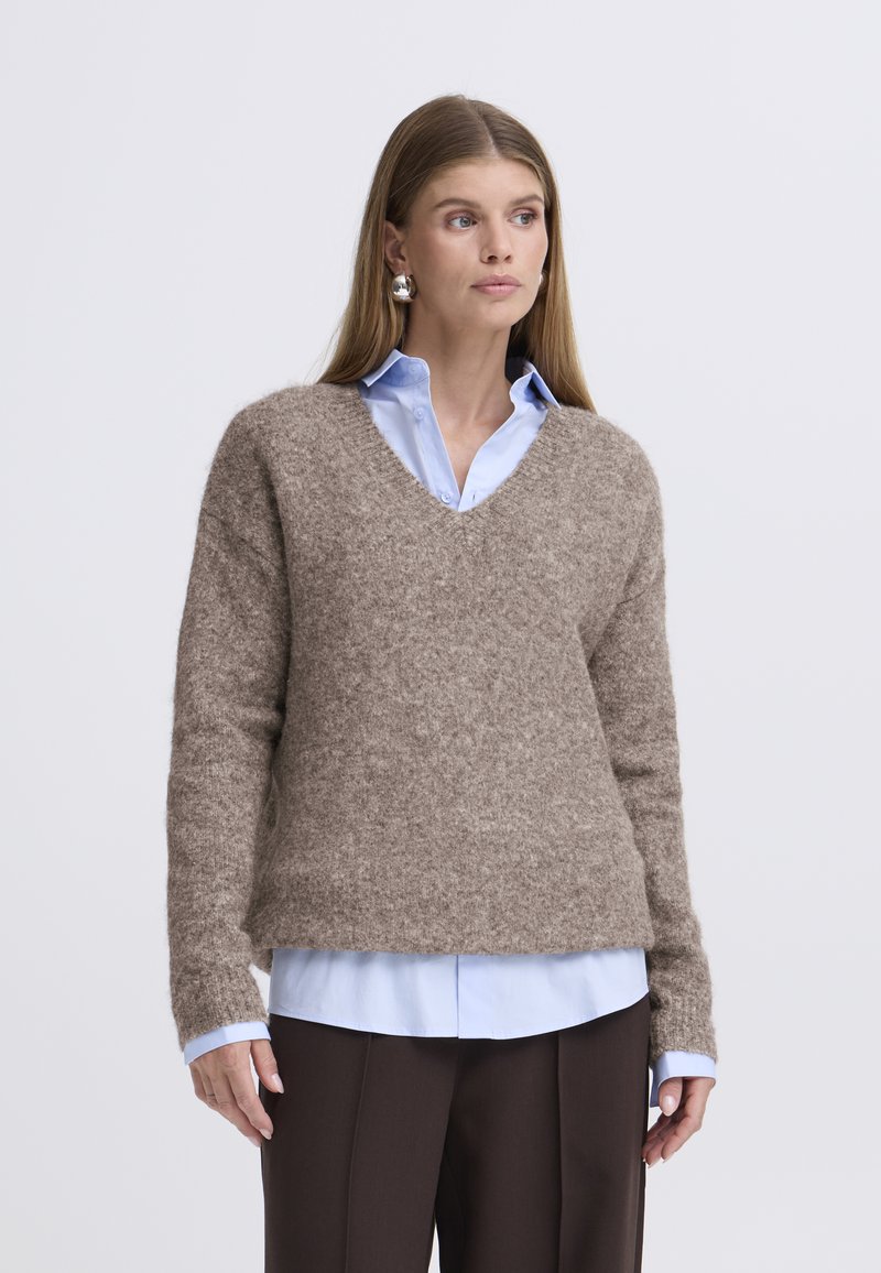 Rjavi, mehak pulover z V-izrezom, dolgimi rokavi in sproščenim krojem, nošen čez svetlo modro srajco z gumbi. Gladek tekstura in priložnostna zasnova.