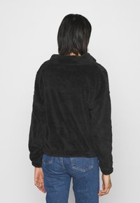 Sudadera negra de forro polar con cuello alto, mangas largas y corte relajado. Textura suave y puños elásticos, combinada con jeans de mezclilla azules.