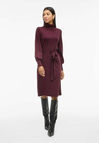 Robe tricotée bordeaux avec un col montant, des manches longues et une taille nouée. Assortie à des bottes noires à hauteur de genou pour un contraste. Texture lisse.