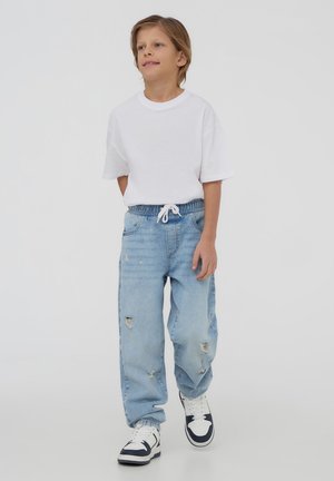 T-shirt bianca in cotone, dal taglio morbido, abbinata a jeans chiari strappati e sneakers con accenti blu navy. Design semplice, outfit casual.