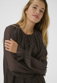 Noor noor blondide juustega noor naine kannab läbipaistvat tumedat pruunist bluus, millel on rikkaid nööpe ja sidumine kaeluses, naeratades õrnalt.