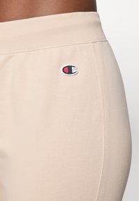 Pantalons de jogging beige avec une texture douce et lisse, une taille élastique et un petit logo brodé en rouge et noir sur le côté.