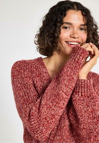 Kvinde med krøllet hår, der smiler, iført en varm rød og hvid strikket sweater, med hænderne nær ansigtet mod en simpel lys baggrund.