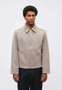 Męska krótka kurtka w beżowym materiale o gładkiej fakturze. Posiada zamek błyskawiczny z przodu, zaostrzony kołnierz i delikatne szwy na ramionach.