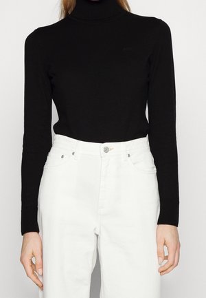 Personne portant un pull noir à col roulé ajusté, rentré dans un jean blanc taille haute avec boutons et poches sur le devant.