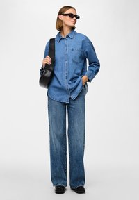 Svetlo modra denim srajca oversized kroja s sprednjim žepom, v kombinaciji s širokimi hlačami. Črna torba za ramo in pravokotna sončna očala dopolnjujeta videz.