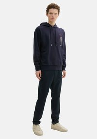Granatowa bluza z kapturem ze sznurkiem, z grafiką na klatce piersiowej. W zestawie z ciemnymi spodniami i jasnymi trampkami.