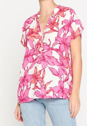 Woman wearing a loose white blouse with bright pink and red floral print, short sleeves, and light blue jeans.