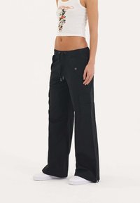 Zwarte cargobroek met een elastische tailleband en trekkoord, voorzakken, zijcargo zak en wijde pijpen. Gepaard met witte schoenen.