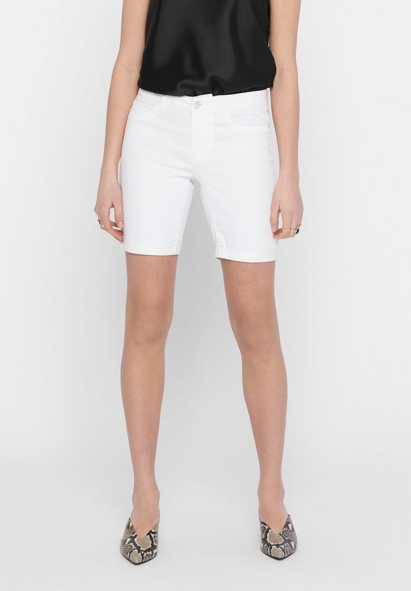 Femme portant un short en denim blanc mi-cuisse, un haut noir sans manches et des talons pointus à motifs, debout devant un fond blanc uni.