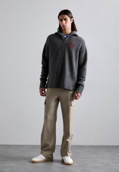 Dunkelgrauer Wollpullover mit einem Reißverschluss am Hals, versehen mit einem roten gestickten Logo. Kombiniert mit beigen Cargo-Hosen und weißen Sneakers.