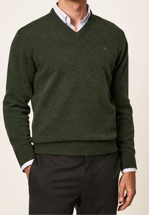 Mężczyzna ubrany w ciemnozielony sweter z dekoltem w serek na białej koszuli oraz czarne spodnie, ręce swobodnie opuszczone, na neutralnym tle.