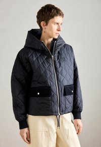 Marineblaue gesteppte Jacke mit Reißverschluss vorne, Kapuze und zwei schwarzen Samt-Taschen. Mit gerippten Manschetten und einem Kordelzugsaum.