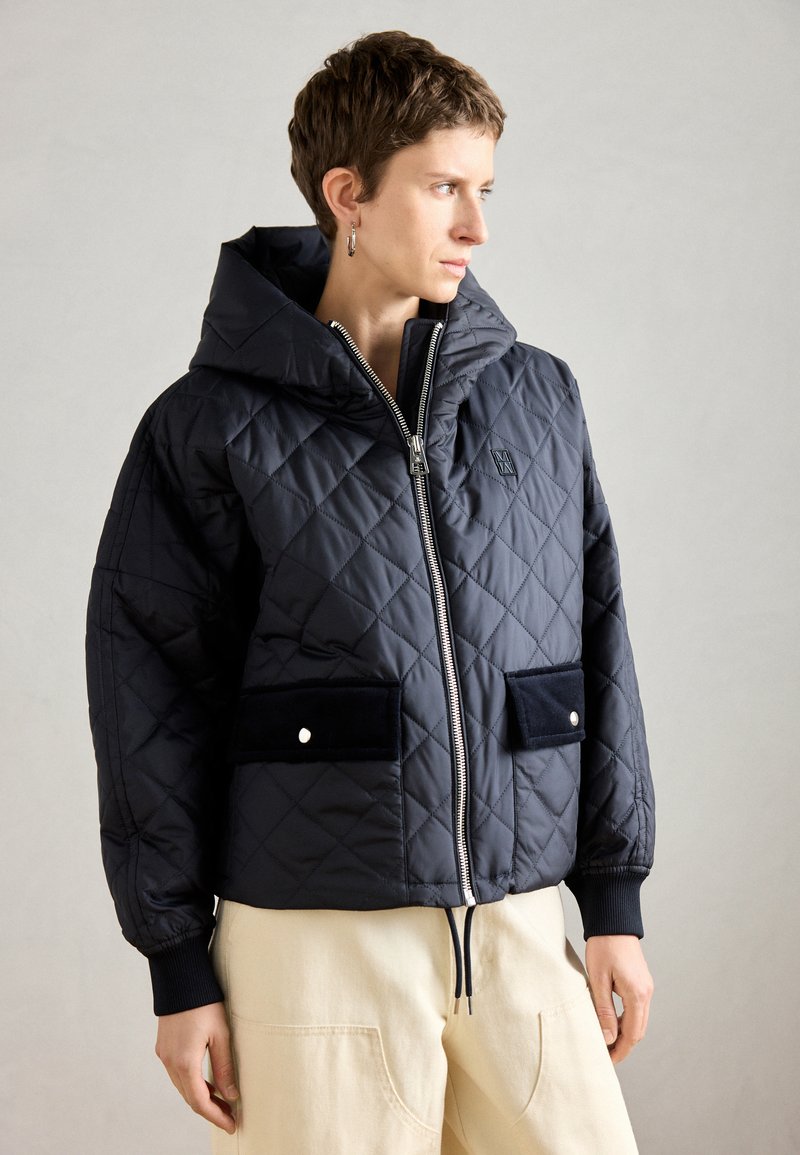 Marineblaue gesteppte Jacke mit Reißverschluss vorne, Kapuze und zwei schwarzen Samt-Taschen. Mit gerippten Manschetten und einem Kordelzugsaum.