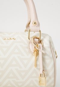 Beige Handtasche mit strukturiertem geometrischen Muster, goldenen Beschlägen und einem Schlüssel-Anhänger. Verfügt über cremefarbene Griffe und metallische Akzente. Markenlogo sichtbar.
