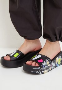 Sandale platformă negre cu talpă joasă, având o curea largă cu accente neon și un design de tip graffiti pe laterale. Textură netedă, stil cu vârful deschis.