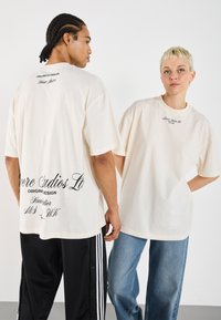 Cream-colored oversized t-shirts with short sleeves feature black text graphics on the back and chest, set against a neutral background.