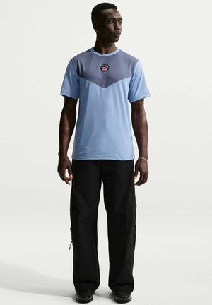 Uomo in piedi, indossa una t-shirt Nike azzurro chiaro e grigia con logo sul petto, pantaloni neri larghi e scarpe nere su uno sfondo semplice.