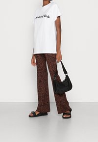 Vit bomullströja med "Nothing Club"-grafik, i kombination med bruna byxor i zebramönster och en svart läderväska; platta svarta sandaler fulländar looken.