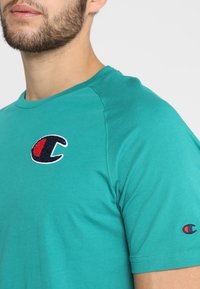 Muž s plnovousem nosící tyrkysovou košili s krátkým rukávem, na které jsou na hrudi a levém rukávu červené a černé loga Champion.