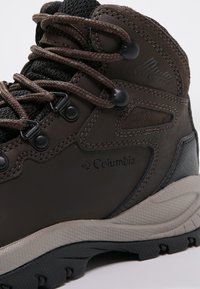 Bota de senderismo impermeable marrón con parte superior texturizada, suela de goma gris y cordones marrones. Presenta un logo y ojales reforzados para mayor durabilidad.
