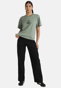 Olive green cotton t-shirt with a round neckline and short sleeves, featuring a black snake graphic on the front; paired with black wide-leg pants.